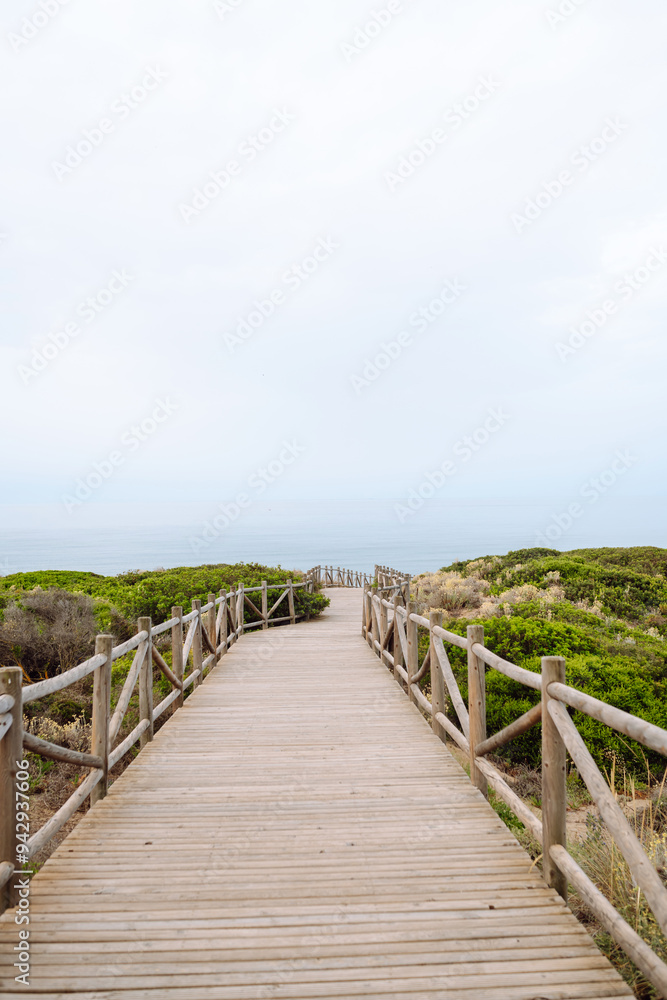 A wooden walkway winds through lush coastal vegetation, directing attention towards the calm sea. The overcast sky adds a serene atmosphere. A wooden walkway winds through lush coastal vegetation, directing attention towards the calm sea. The overcast sky adds a serene atmosphere.