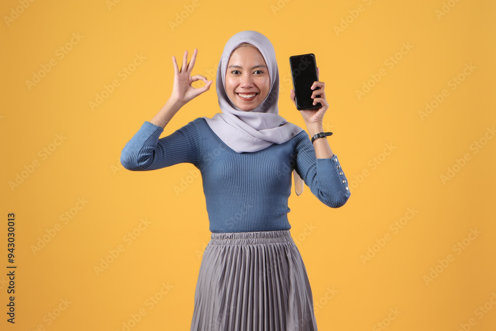 happy asian indonesian muslim woman holding smart phone and showing smart phone blank screen and giving ok finger gesture on isolated yellow background happy asian indonesian muslim woman holding smart phone and showing smart phone blank screen and giving ok finger gesture on isolated yellow background