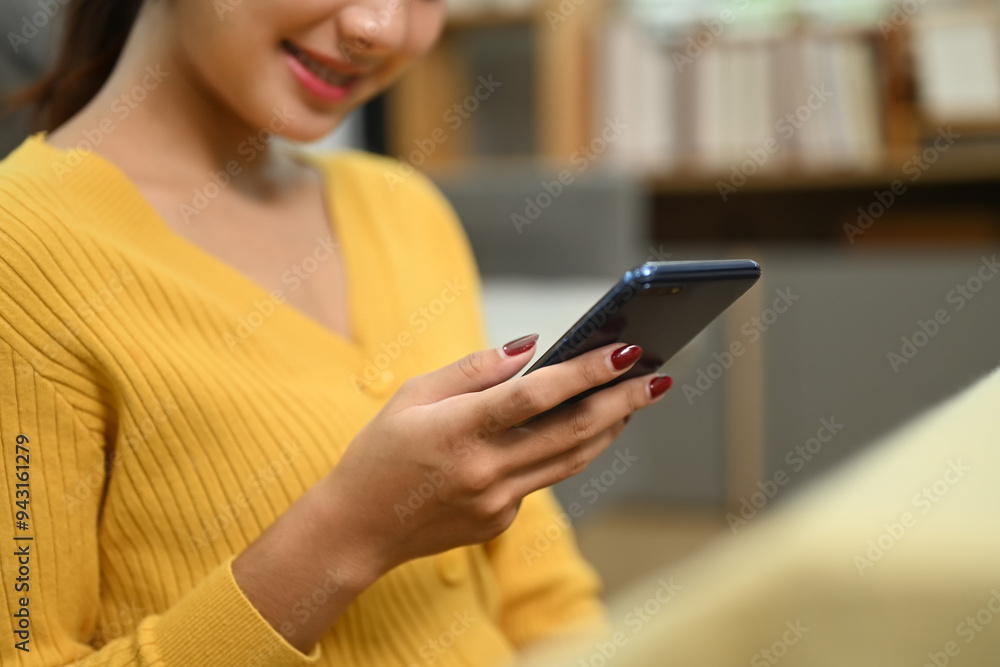 Cropped shot smiling young woman in yellow cardigan using mobile phone in living room Cropped shot smiling young woman in yellow cardigan using mobile phone in living room