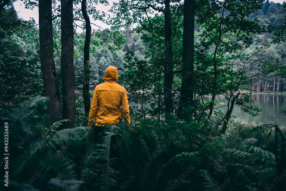 Young man poses wearing his yellow coat and hat among green leaves. He walks in the forest and enjoys nature. Young man poses wearing his yellow coat and hat among green leaves. He walks in the forest and enjoys nature.