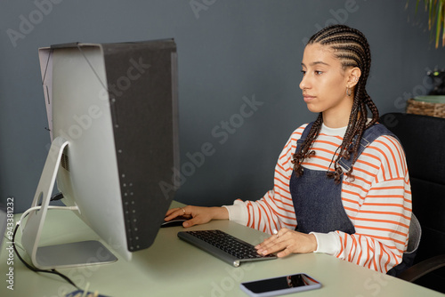 Minimal side view portrait of young Black woman with cornrows hairstyle using computer while editing video or pictures in office photo