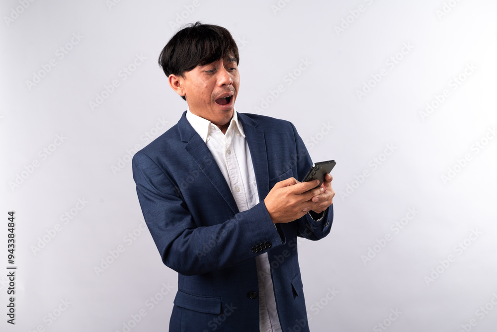 A surprised Asian businessman in a navy blue suit looks at his smartphone with a shocked expression. He holds the phone with both hands, unexpected notification. White background. A surprised Asian businessman in a navy blue suit looks at his smartphone with a shocked expression. He holds the phone with both hands, unexpected notification. White background.