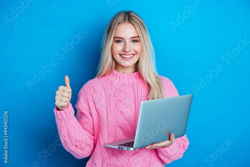 The portrait of a nice young woman thumbing up the laptop while wearing a pink sweater is isolated on a blue color background