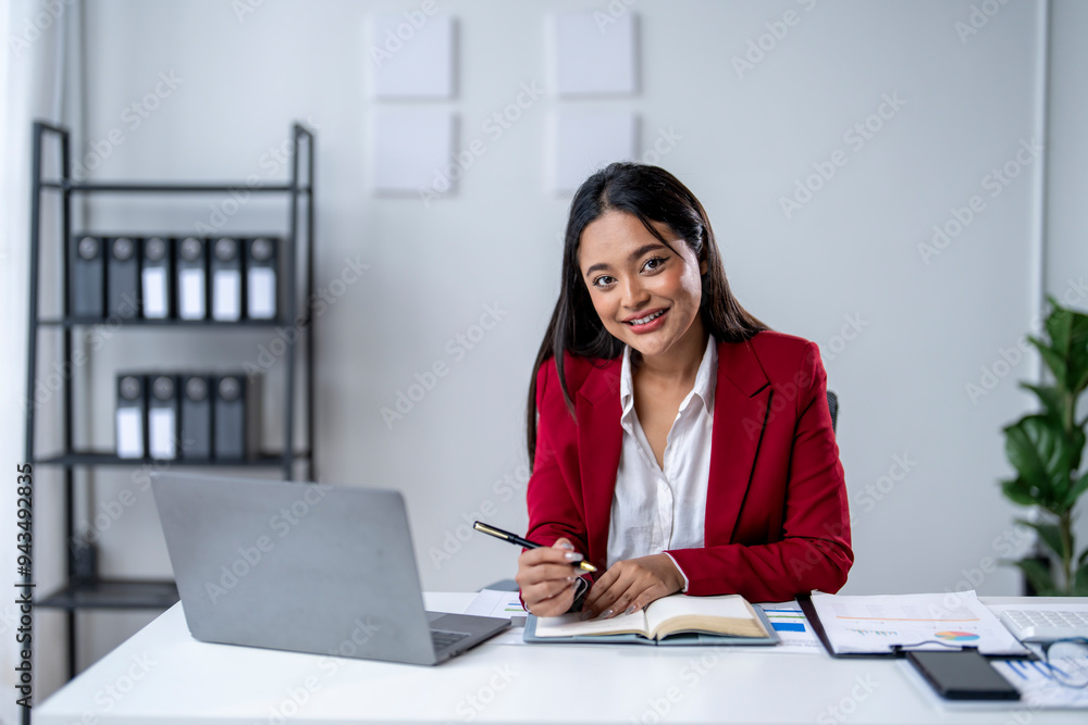 A woman in a red jacket is sitting at a desk with a laptop and a notebook A woman in a red jacket is sitting at a desk with a laptop and a notebook