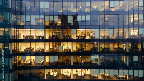Drone shot offers a panoramic view a towering glass skyscraper, where office lights reveal business professionals continuing their tasks late into the night, reflecting the nonstop pace of city life.