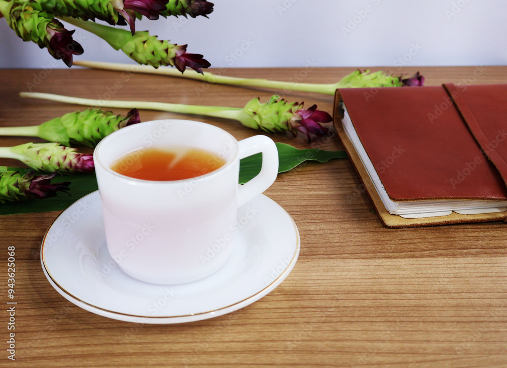 Red-brown hot tea in a ceramic cup and saucer sits between books and flowers on a wide wooden table.
Red-brown hot tea in a ceramic cup and saucer sits between books and flowers on a wide wooden table.