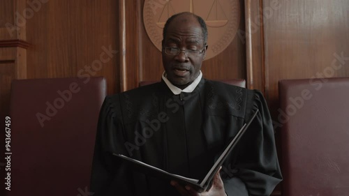 African American male judge dressed in black robe passing verdict while standing with open folder and looking at juridical document during court trial