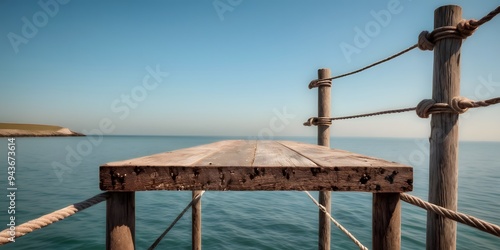 A weathered metal plate mounted on a pole at the end of a wooden quay bridge, overlooking the calm sea under a clear sky.