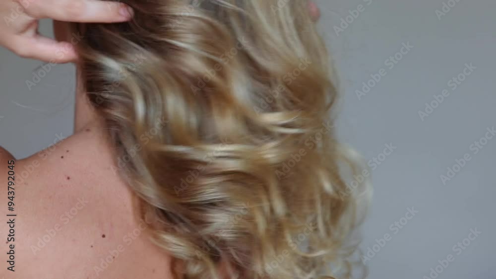 A young blonde woman standing with her back demonstrating her golden wavy hair lifting the curls with hands on a grey background. Result of coloring, highlighting, perming. Beauty and fashion A young blonde woman standing with her back demonstrating her golden wavy hair lifting the curls with hands on a grey background. Result of coloring, highlighting, perming. Beauty and fashion