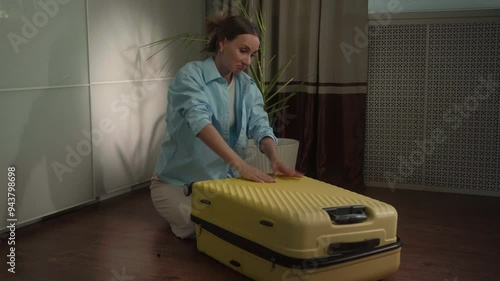 Woman struggles to close her overpacked yellow suitcase while preparing for a trip. Pushing down hard, she battles the challenge of fitting everything inside photo