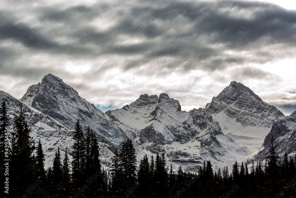 Dramatic dark clouds above the snow covered mountain peaks of the Three Sisters with silhouetted evergreen trees in the foreground; South of Canmore, Alberta, Canada Dramatic dark clouds above the snow covered mountain peaks of the Three Sisters with silhouetted evergreen trees in the foreground; South of Canmore, Alberta, Canada