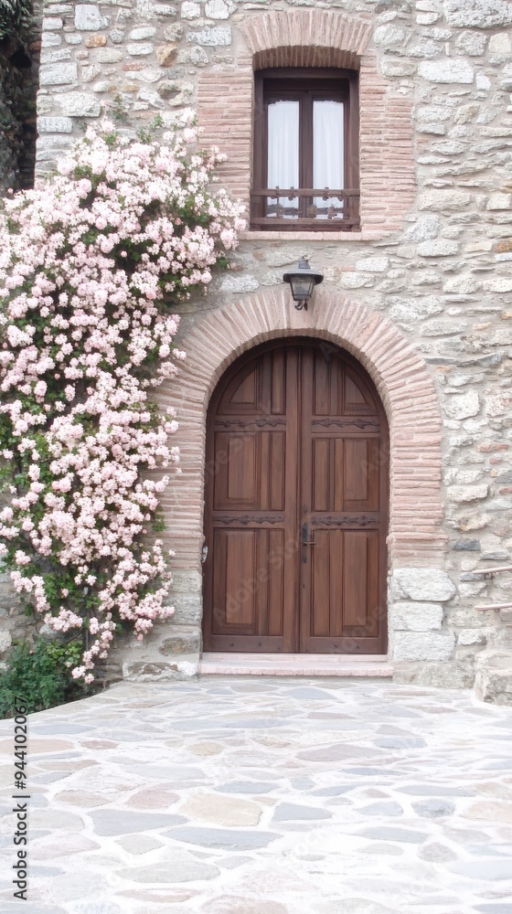 A charming rose bush with pink flowers cascades over a weathered stone wall, framing a small window and door in a peaceful setting in France A charming rose bush with pink flowers cascades over a weathered stone wall, framing a small window and door in a peaceful setting in France