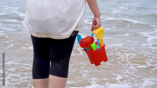 Slow motion shot Indian woman in white shirt standing on sea shore, beach looking at waves, holding a colorful sand castle making kit toy at baga beach in goa India photo