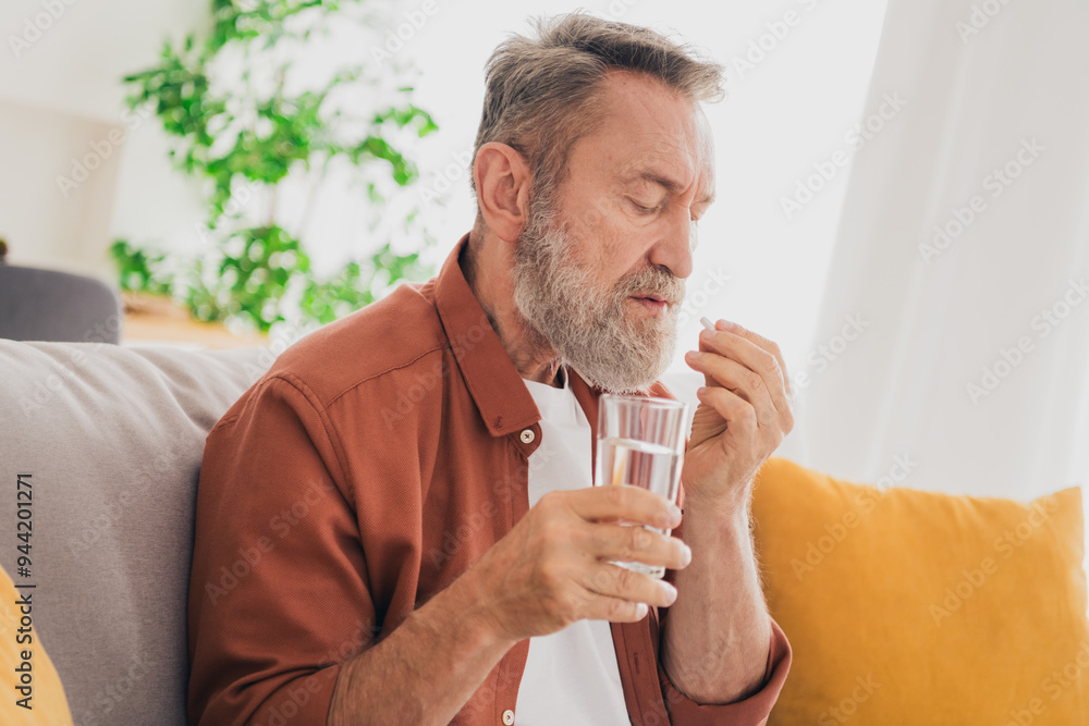 Photo of senior male sit couch take meds drink water glass dressed brown outfit cozy day light home interior living room Photo of senior male sit couch take meds drink water glass dressed brown outfit cozy day light home interior living room