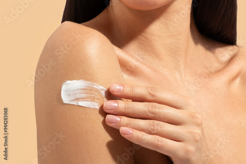 Woman spreading cream on her shoulder, taking care of herself, swatch of cream, beige background, closeup shot photo