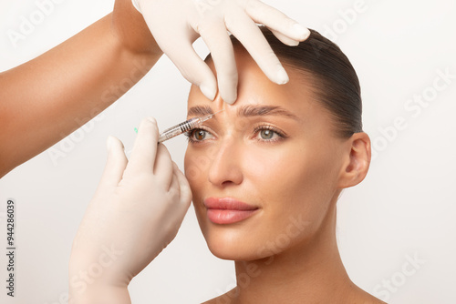 Beautician making hyaluronic acid injection for glabella facial rejuvenation procedure, woman getting cosmetic injection between eyebrows over white studio background photo