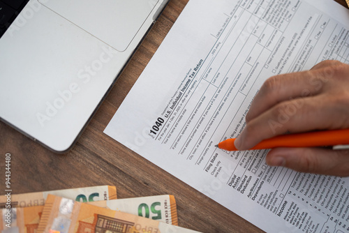Close-up of a person filling out a 1040 tax form with an orange pen next to a laptop. Tax season, personal finance, and accounting concept 