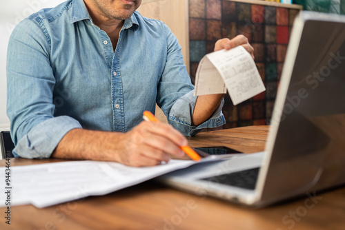 Middle-aged man calculating taxes at a wooden desk with a laptop and holding receipts. Personal finance management and accounting concept during tax season  photo