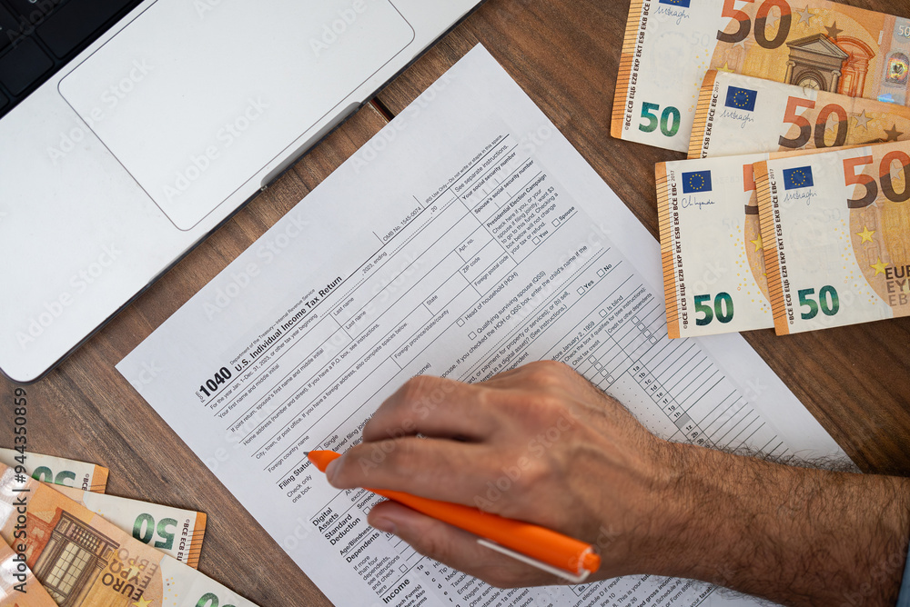 Fototapeta premium Hand of a man filling out a 1040 tax form with euros and a laptop on a wooden desk. Tax preparation and financial documentation concept, suitable for accounting and international finance themes 