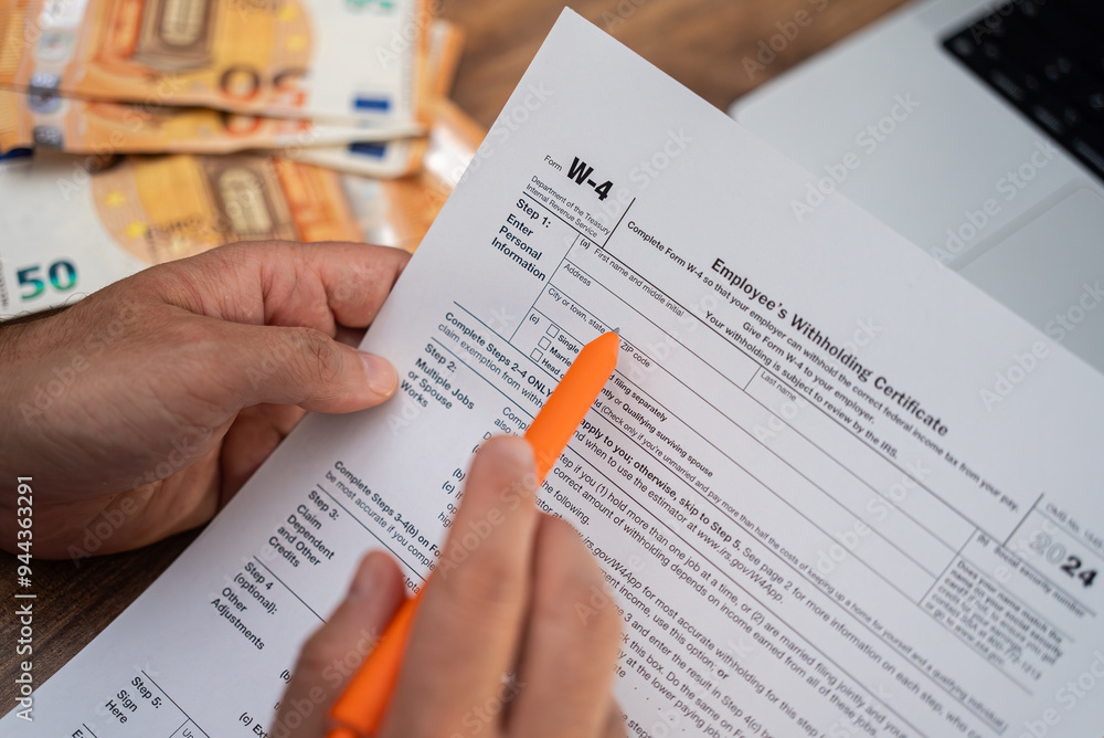 Freelancer meticulously reads W-4 form for employer to know exactly sum of money federal tax to withhold from earnings at dining table with laptop and euro bills Freelancer meticulously reads W-4 form for employer to know exactly sum of money federal tax to withhold from earnings at dining table with laptop and euro bills