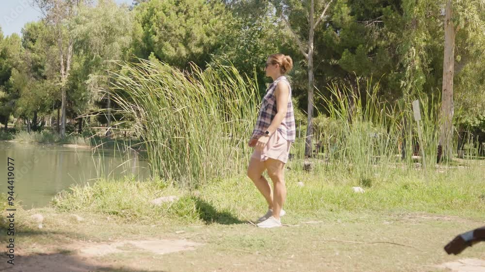 Woman strolls beside a Pond with Tall Reeds, enjoying the Tranquil Park Setting on a Sunny Day. Slow motion. Woman strolls beside a Pond with Tall Reeds, enjoying the Tranquil Park Setting on a Sunny Day. Slow motion.
