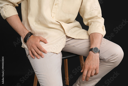 Stylish handsome young man with wristwatch sitting on chair against black background, closeup