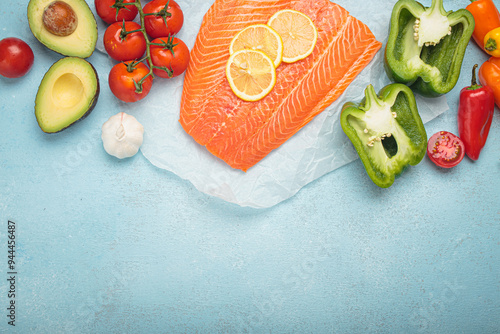 Fresh uncooked salmon fillet seasoned with lemon slices is lying on white baking paper, surrounded by fresh vegetables on a blue background, copy space