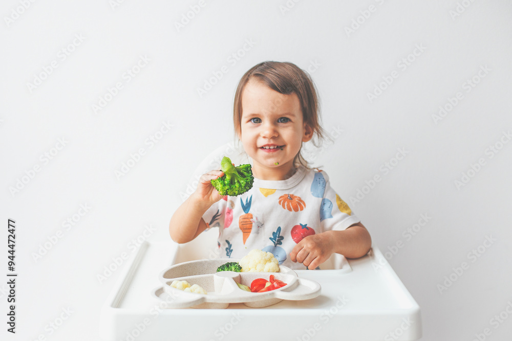 cute girl 1.5 years old sits in a high chair on a white background with a plate of vegetables, holds broccoli in her hand, smiles and looks at the camera cute girl 1.5 years old sits in a high chair on a white background with a plate of vegetables, holds broccoli in her hand, smiles and looks at the camera