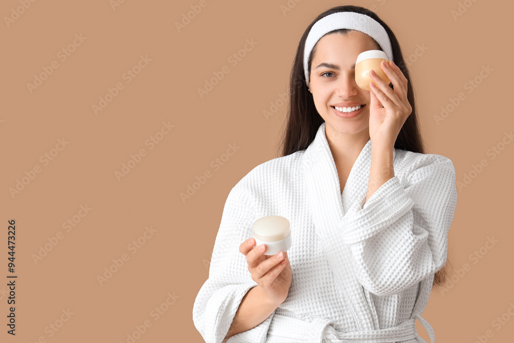 Beautiful young happy woman in bathrobe with jars of cream on beige background Beautiful young happy woman in bathrobe with jars of cream on beige background