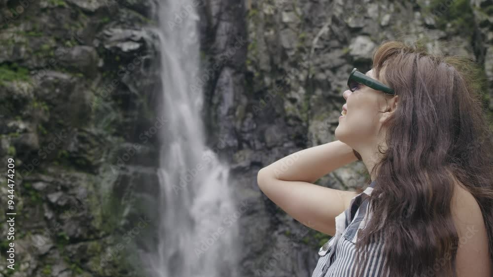 Portrait of a beautiful young woman with brunette hair and wearing sunglasses, resting against a backdrop of a large waterfall. The woman is smiling affably and is facing the camera. Slow motion.