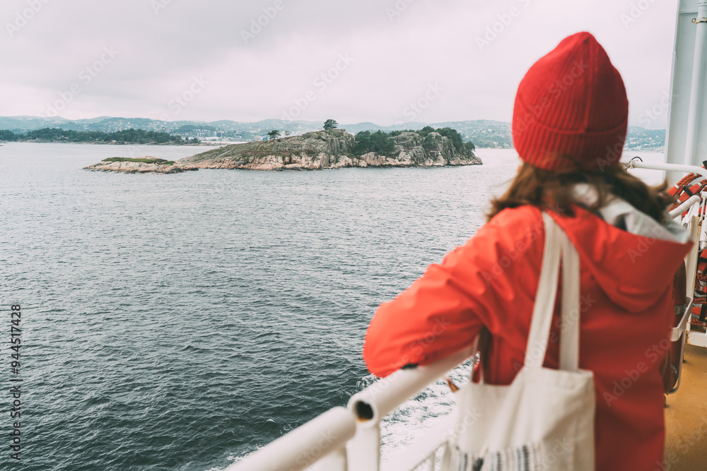 Woman dressed in red jacket and beanie, stands on Norwegian ferry's deck, gazing out at small rocky island in distance. Scene captures serenity of Nordic travel under overcast skies Woman dressed in red jacket and beanie, stands on Norwegian ferry's deck, gazing out at small rocky island in distance. Scene captures serenity of Nordic travel under overcast skies