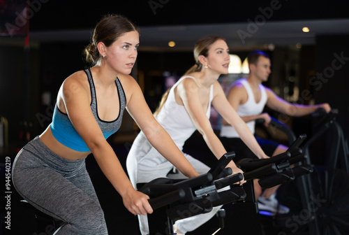 Young woman spends time in gym, improves her physical shape with exercises. Female sport studio hall visitor uses exercise bike to pump leg muscles. Strength and motivation, sport, fitness goal