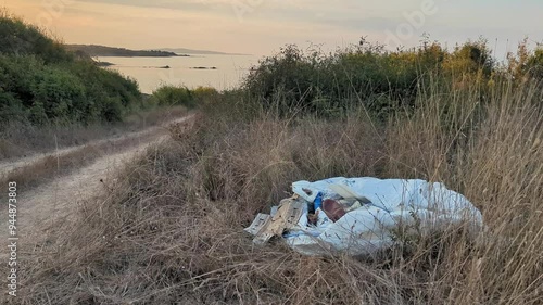 A bag of mixed household trash discarded in nature, near a dirt road with a view of the sea. Highlights the problem of people's disregard for nature and illegal environmental pollution. photo