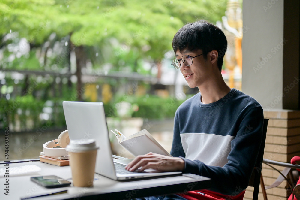 A young, positive Asian male college student is using his laptop and researching in a book. A young, positive Asian male college student is using his laptop and researching in a book.