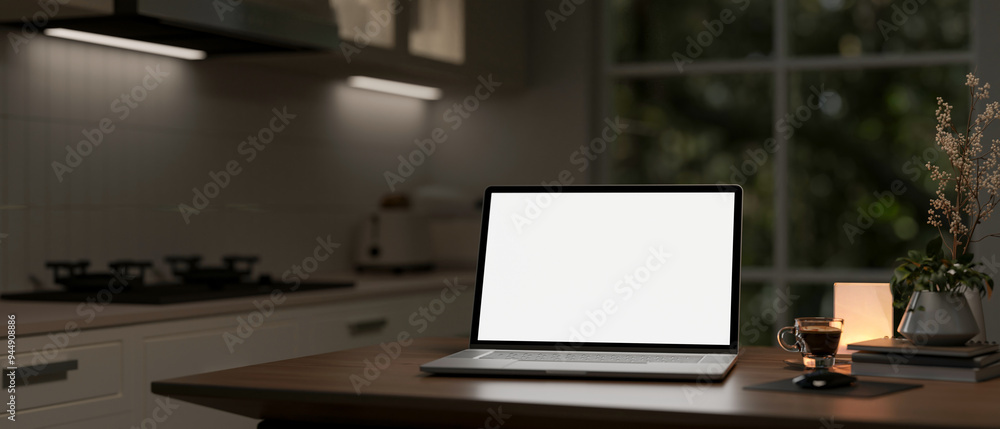 A laptop computer with a white screen mockup on a wooden table in a modern kitchen at nighttime.