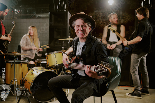 Selective focus portrait of joyful mature Caucasian bassist sitting on chair holding guitar smiling at camera during rehearsal with his band