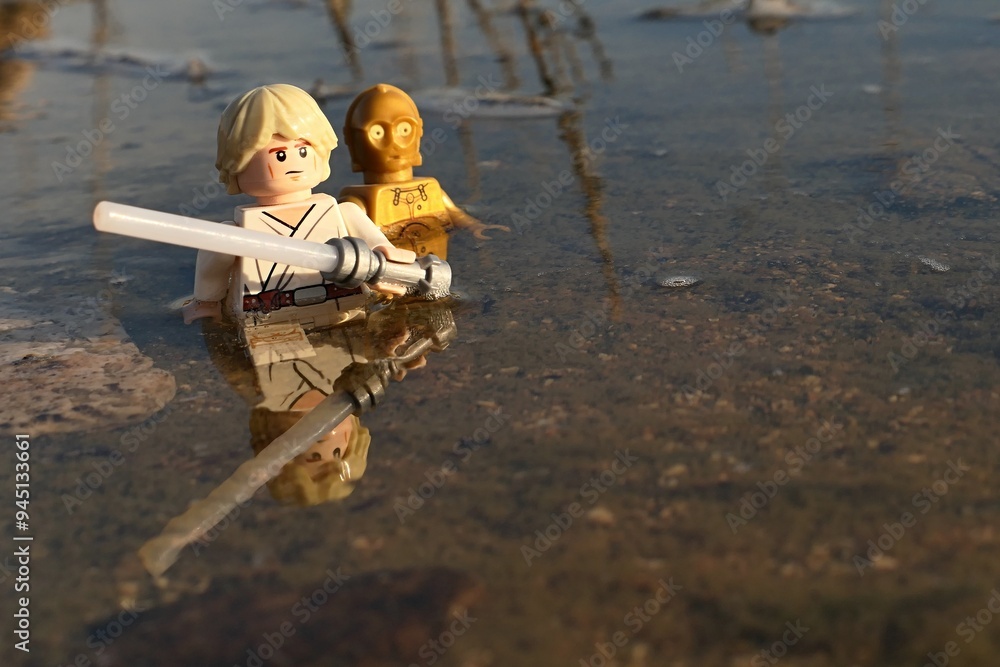 Naklejka premium LEGO Star Wars figures of jedi knight Luke Skywalker with white lightsaber and golden android C3PO walking waist deep through waters of shoreline marsh.