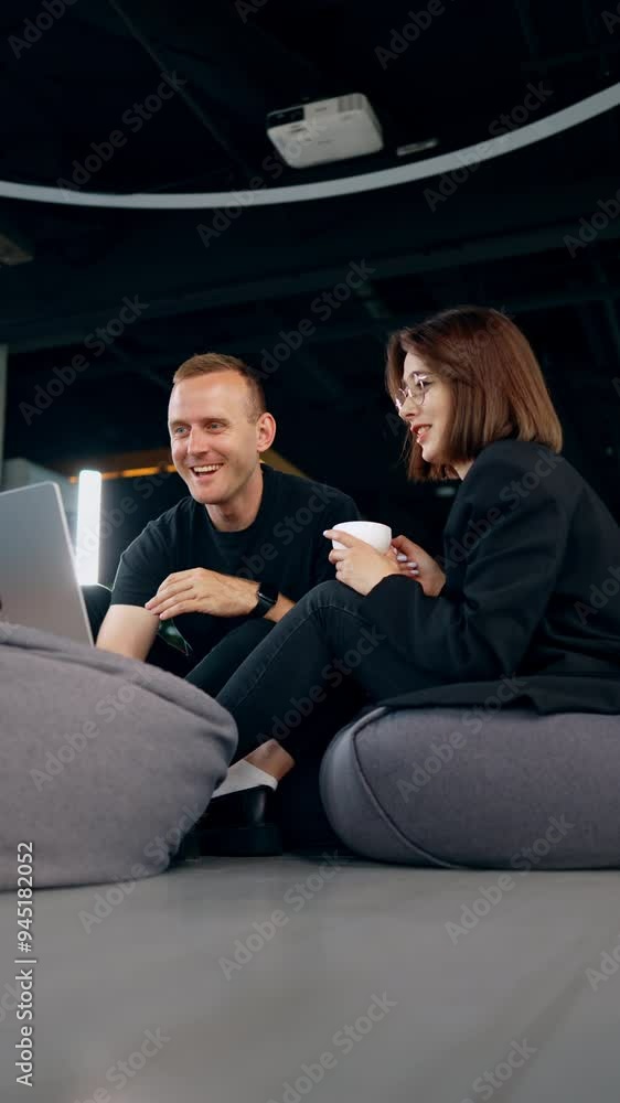 Young male and female co-workers sit in bean bag chairs communicating cheerfully. Friendly colleagues discuss video on laptop. Low angle view. Vertical video. Young male and female co-workers sit in bean bag chairs communicating cheerfully. Friendly colleagues discuss video on laptop. Low angle view. Vertical video.