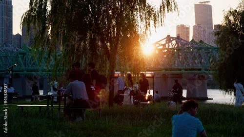People Relaxing at Nodeul Island Park by the Han River in Seoul, South Korea During Golden Hour with Yeouido Skyline and Railway Bridge in the Background
