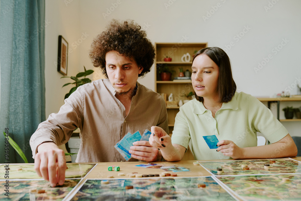 Fototapeta premium Young gen Y biracial couple sitting at table in living room talking while playing modern fantasy board game