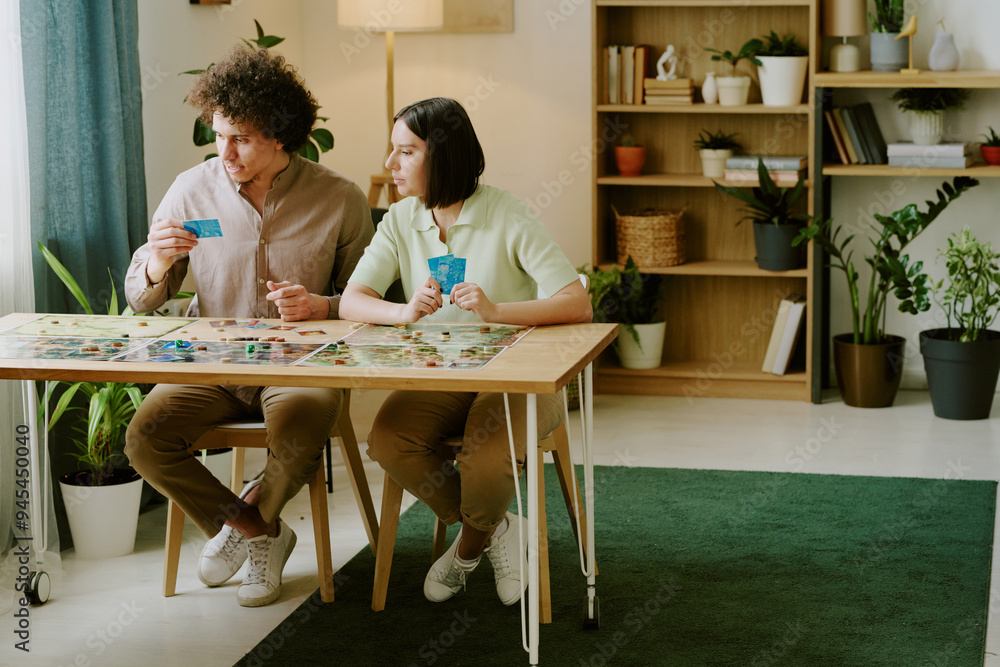 Modern gen Y biracial man and woman sitting at wooden table in living room at home playing fantasy board game Modern gen Y biracial man and woman sitting at wooden table in living room at home playing fantasy board game