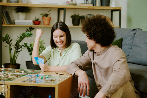 Cheerful young Caucasian woman sitting at coffee table playing board game with her boyfriend and winning round photo