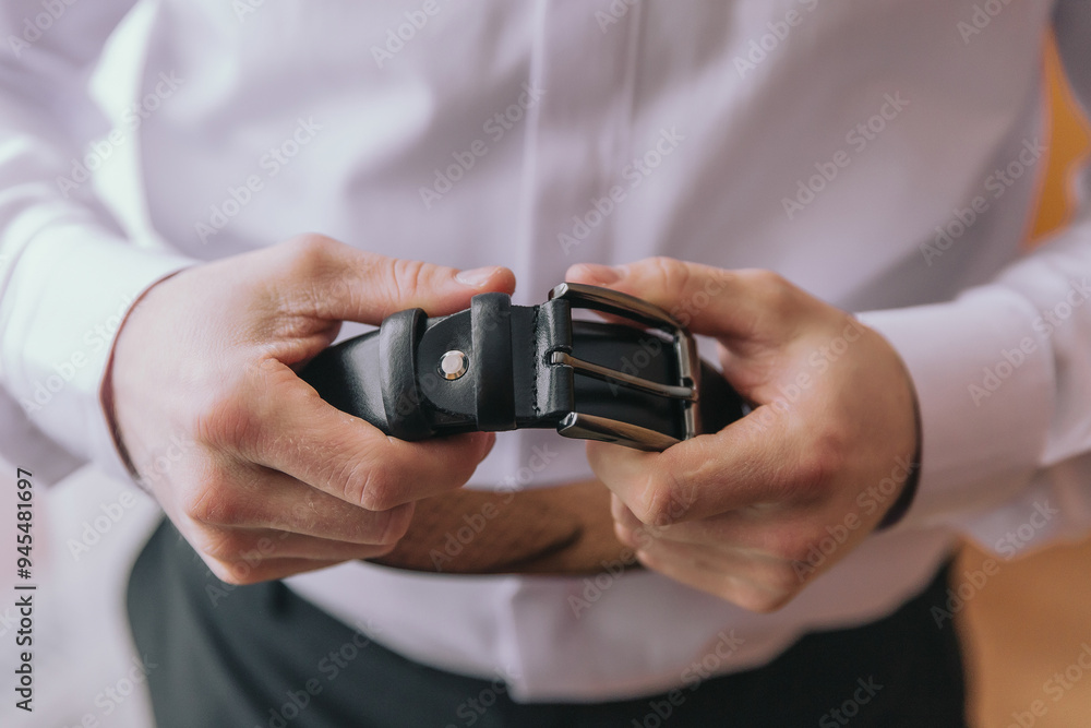 A man is holding a belt in his hands. The belt is black and has a silver buckle. The man is wearing a white shirt and black pants A man is holding a belt in his hands. The belt is black and has a silver buckle. The man is wearing a white shirt and black pants