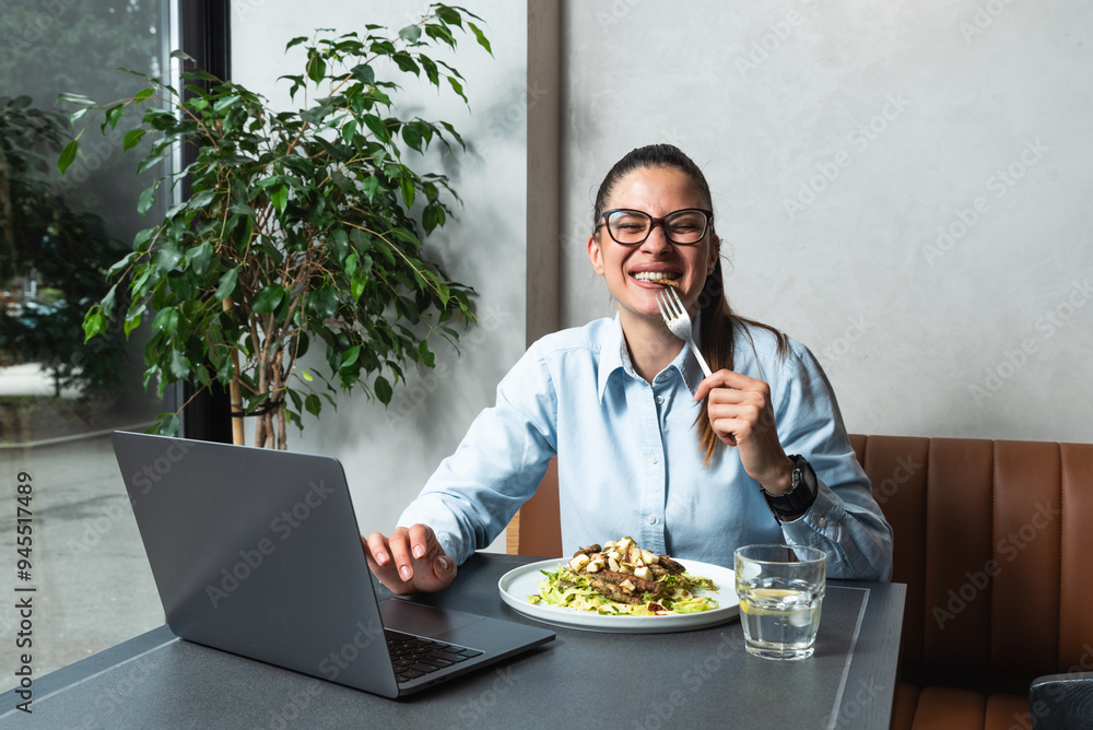 Young business woman working remotely from cafeteria during lunch break. Female office worker using laptop computer while she eating at restaurant to work or watching funny content on internet. Young business woman working remotely from cafeteria during lunch break. Female office worker using laptop computer while she eating at restaurant to work or watching funny content on internet.