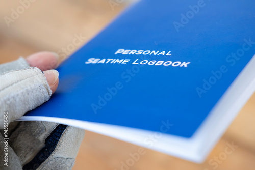 Gloved hand holding blue Personal Seatime Logbook. Maritime record keeping. Seafarer documentation. Nautical career tracking, Seafarer's chronicle, Sea hours documented photo