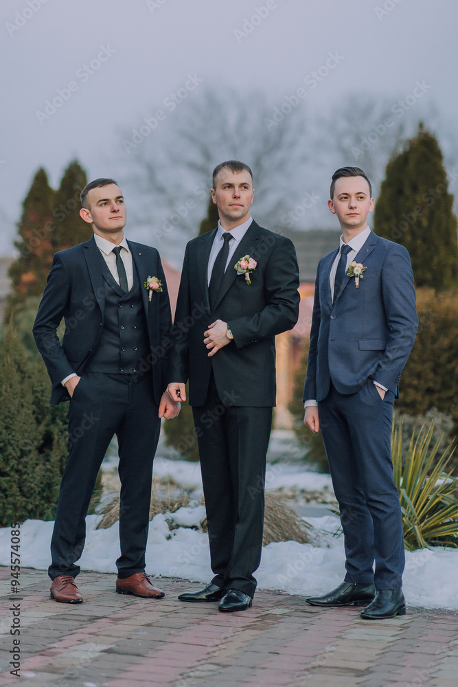 Three men in suits stand together in a snowy courtyard. They are all wearing ties and have flowers in their lapels. Scene is formal and elegant, as the men are dressed in suits Three men in suits stand together in a snowy courtyard. They are all wearing ties and have flowers in their lapels. Scene is formal and elegant, as the men are dressed in suits