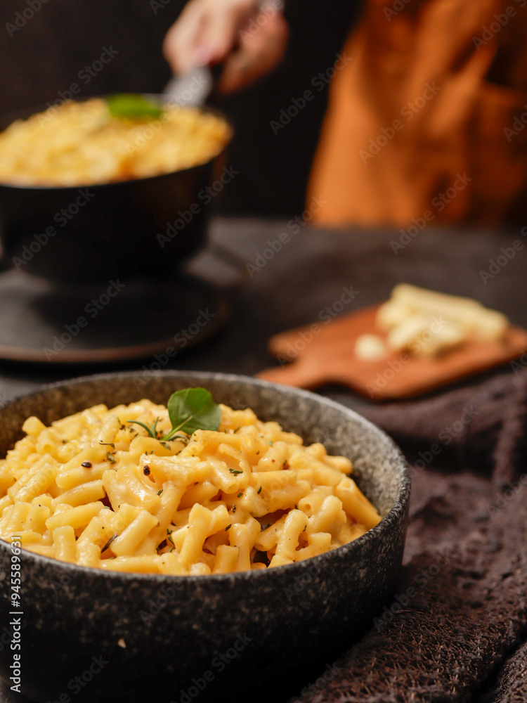A black bowl filled with creamy macaroni and cheese, garnished with fresh basil leaves, sits on a dark surface. A blurred hand in the background is holding another bowl of the same dish.