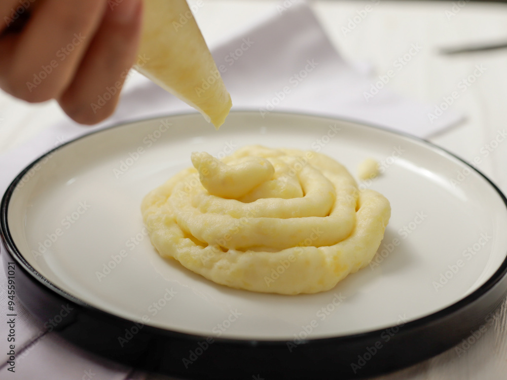 Close-up of a hand squeezing mashed potatoes from a piping bag onto a white plate. The creamy potatoes are smooth and perfectly shaped, with a background of soft white linen. Close-up of a hand squeezing mashed potatoes from a piping bag onto a white plate. The creamy potatoes are smooth and perfectly shaped, with a background of soft white linen.