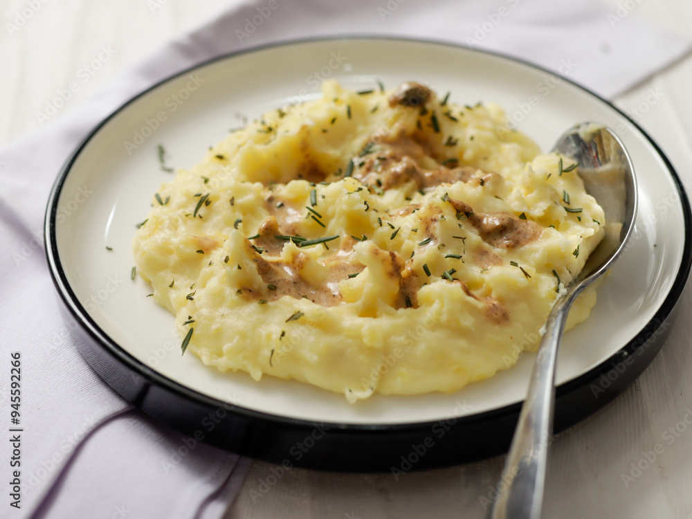 A plate of creamy mashed potatoes topped with a rich, savory gravy and garnished with fresh herbs. The dish is served on a white plate, with a spoon placed on the side, ready to be enjoyed A plate of creamy mashed potatoes topped with a rich, savory gravy and garnished with fresh herbs. The dish is served on a white plate, with a spoon placed on the side, ready to be enjoyed