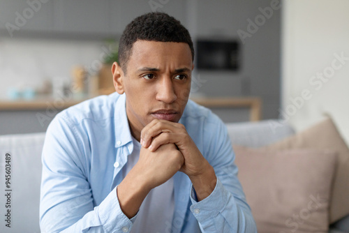 Troubled African American guy looking at copy space while thinking about something, sitting on couch, leaning on his hands, having problem